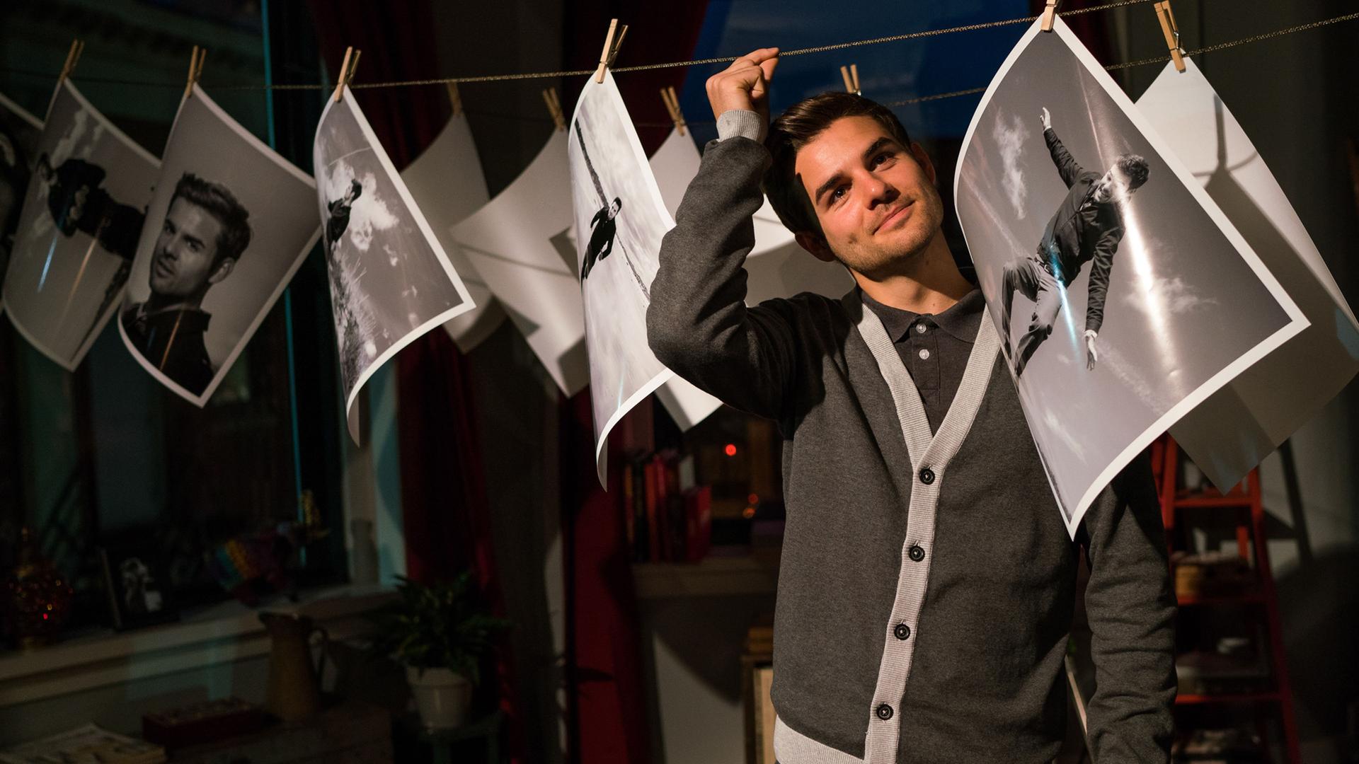 Anthony Cahill (Lucas Reiber) findet es lustig, dass er auf allen Fotos seiner Schwester Lauren ist. Er steht in ihrer Wohnung inmitten zahlreicher großer Fotoabzüge, die an einer Leine hängen.