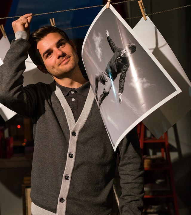 Anthony Cahill (Lucas Reiber) findet es lustig, dass er auf allen Fotos seiner Schwester Lauren ist. Er steht in ihrer Wohnung inmitten zahlreicher großer Fotoabzüge, die an einer Leine hängen.
