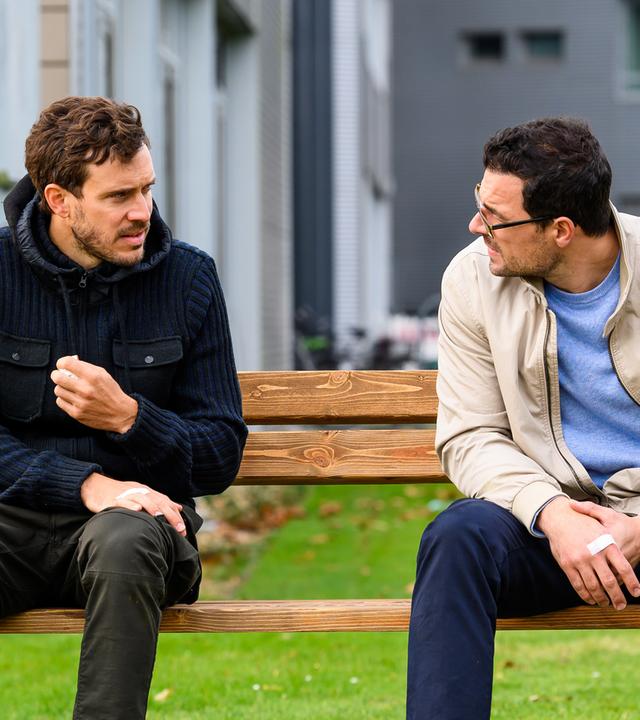 Die Brüder Mirko Koopmann (Tobias van Dieken) und Dr. Tom Koopmann (Tobias Licht) sitzen vor der Klinik auf einer Parkbank. Beide haben Pflaster auf ihren Händen.