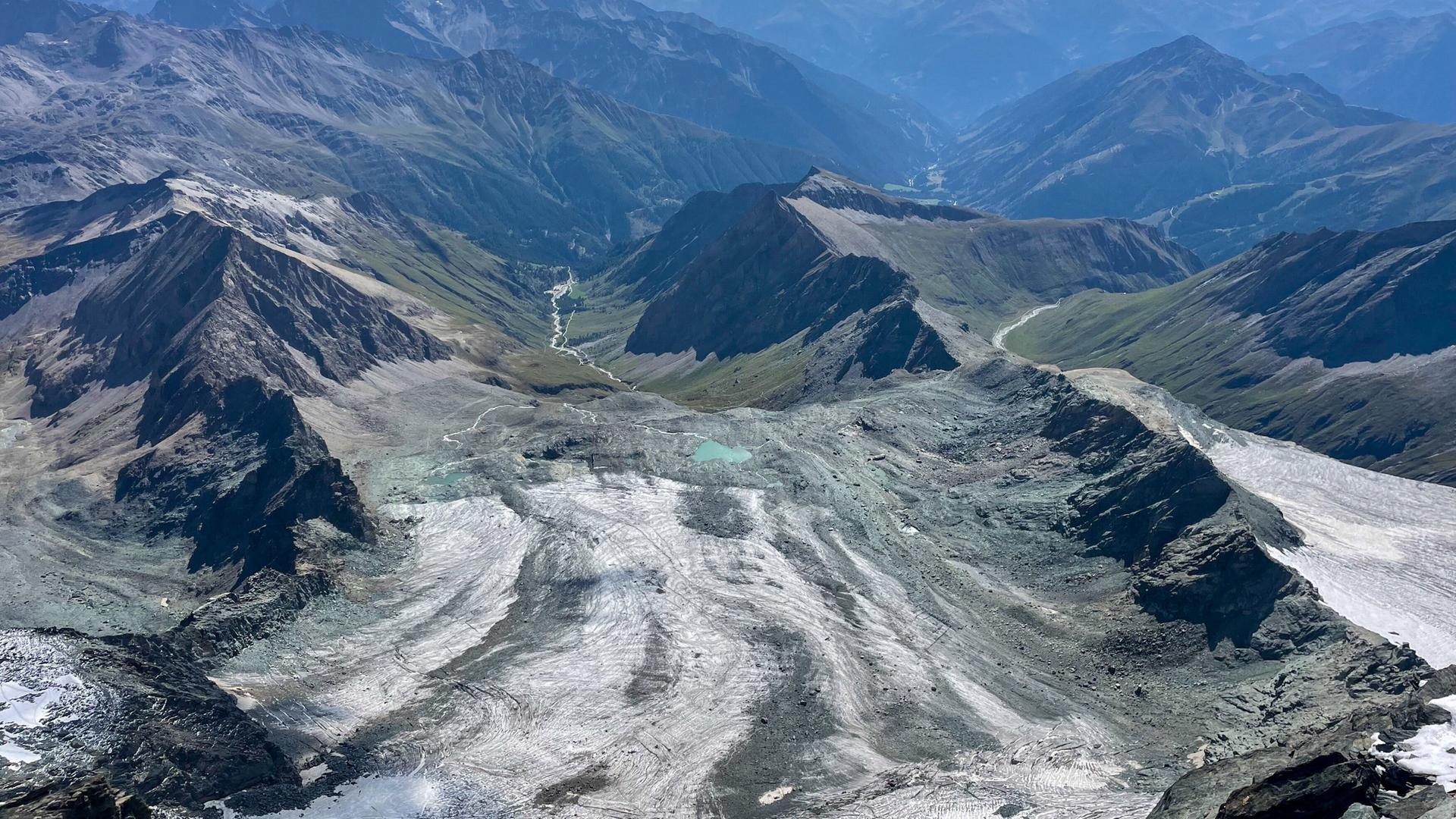 Das Bild zeigt eine atemberaubende Berglandschaft in den Alpen. Im Vordergrund sind große, graue Gletscherflächen sichtbar, die sich in Wellen über das Terrain ausbreiten. Diese Gletscher zeigen deutliche Zeichen des Rückgangs, mit großflächigen, schimmernden Eisschichten, die sich von den Felsen abheben.  Die Umgebung ist geprägt von zerklüfteten Berghängen und schroffen Gipfeln, die in der Ferne in sanfteren grünen Hügeln übergehen. Mehrere scharfe, dunkle Felskanten sind im Vordergrund zu sehen, während die Höhenlagen hinter den Gletschern sich in sanften Blautönen und Wolken verlieren. Der Himmel ist klar, und einige Wolken schweben über den Bergen. Eine Wasserstraße schlängelt sich durch die Landschaft, was auf einen möglichen Fluss oder Bach hindeutet.  Diese Szene vermittelt eine eindrückliche Vorstellung von der Schönheit der Natur und der fragilen Situation in den Alpen, insbesondere in Bezug auf den Klimawandel.