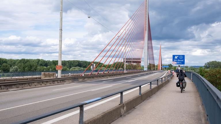 Auf dem Bild sieht man die Bonner Rheinbrücke A565. Die Fahrbahn ist leer und rechts auf dem Bürgersteig rechts fahren zwei Fahrradfahrer:innen.