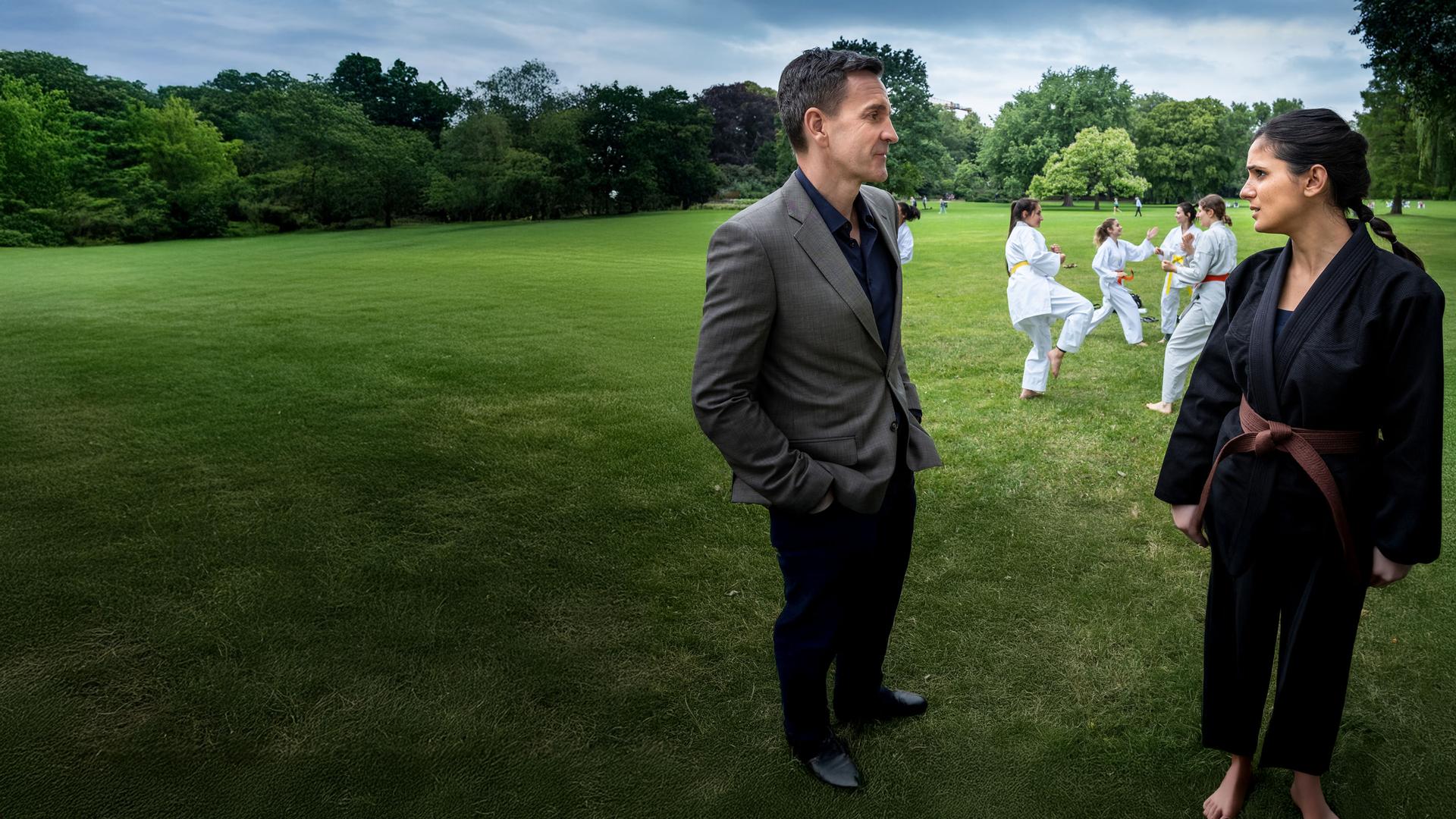 "Notruf Hafenkante - Brüderchen und Schwesterchen": Pinar (Aybi Era) steht im schwarzen Karate-Anzug auf einer Wiese dem LKA-Ermittler Gerald Renner (Michael Ehnert) gegenüber. Sie sprechen miteinander. Im Hintergrund üben jungen Frauen in weißen Anzügen Karate miteinander.