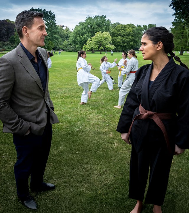 "Notruf Hafenkante - Brüderchen und Schwesterchen": Pinar (Aybi Era) steht im schwarzen Karate-Anzug auf einer Wiese dem LKA-Ermittler Gerald Renner (Michael Ehnert) gegenüber. Sie sprechen miteinander. Im Hintergrund üben jungen Frauen in weißen Anzügen Karate miteinander.