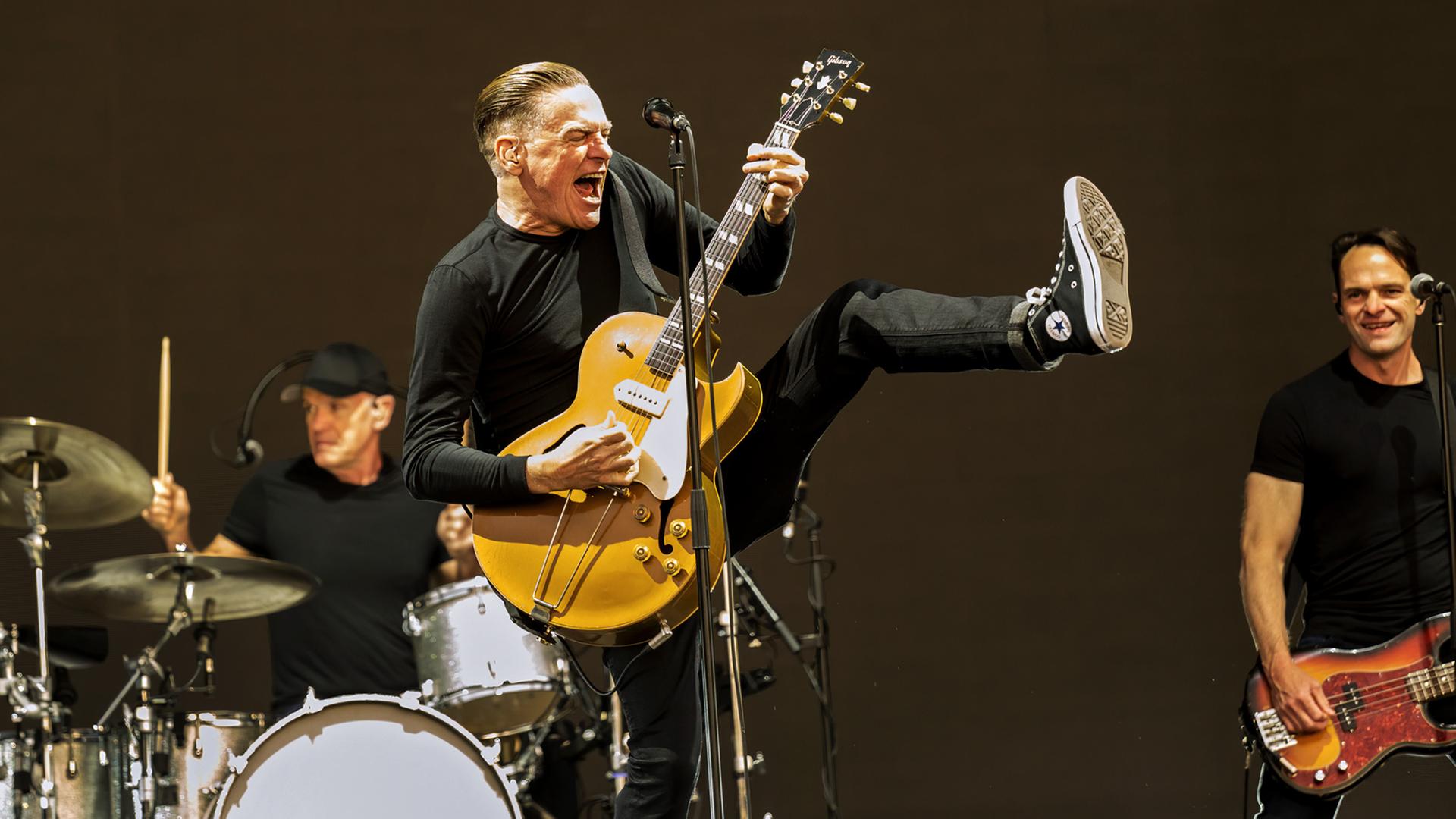 Bryan Adams singt mit seiner Gitarre auf der Bühne. Hinter ihm ist seine Band zu sehen.