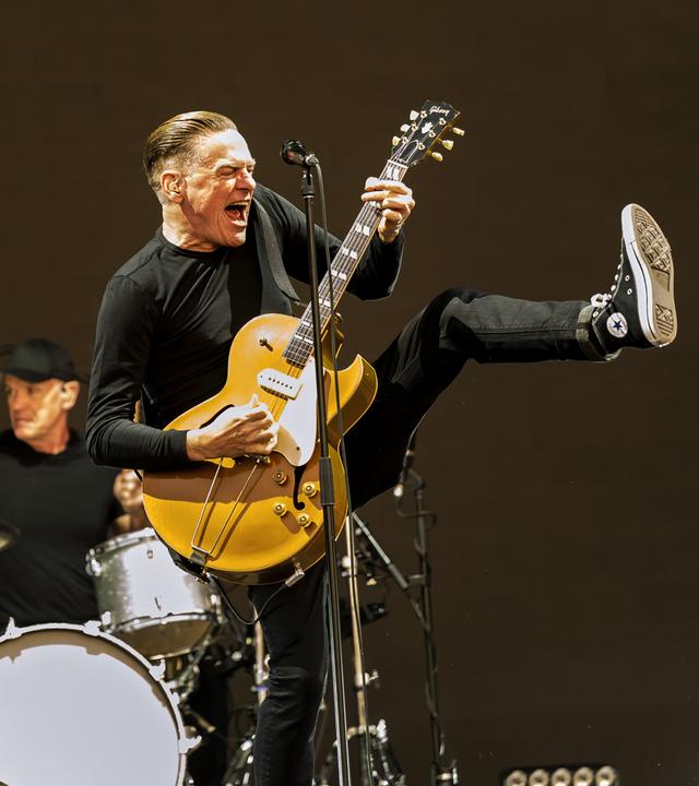 Bryan Adams singt mit seiner Gitarre auf der Bühne. Hinter ihm ist seine Band zu sehen.