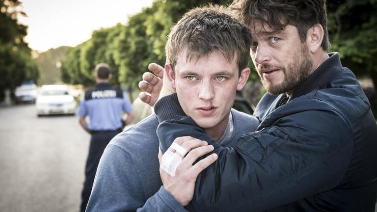 Georg Dengler (Ronald Zehrfeld) steht mit seinem Sohn Jakob (Jannis Niew�hner) in der Zufahrt vom Anwesen des Unternehmers Osterhannes. Georg hat seinen Sohn im Arm. Im Hintergrund sind Polizeiautos und sowie ein Polizeibeamter zu sehen.