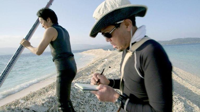 Eroberung der Weltmeere (ARTE, 4.6.2016): Eine japanische Wissenschaftlerin und ein japanischer Wissenschaftler stehen auf einem schmalen Damm im Meer und führen Vermessungsarbeiten durch.