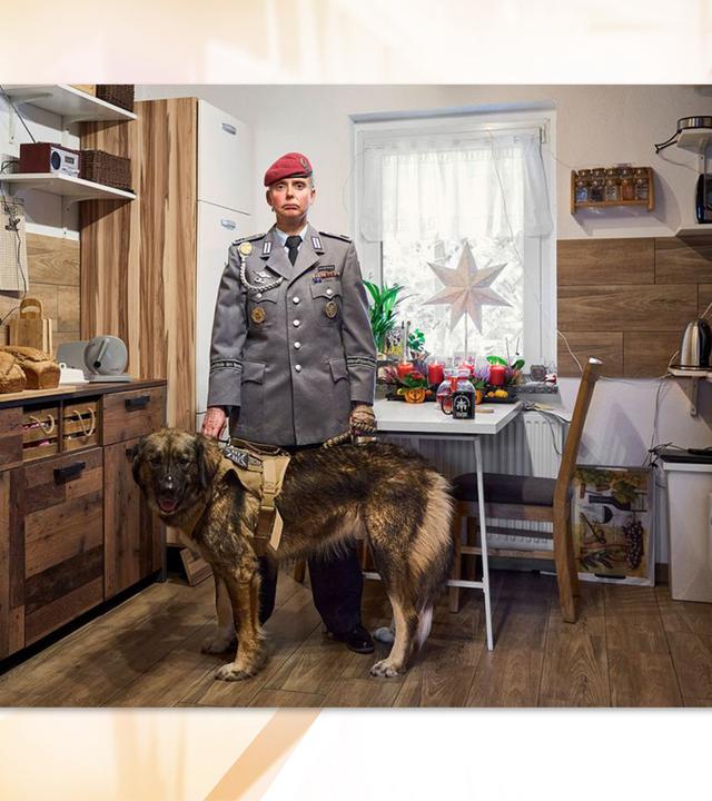 Man sieht eine Soldatin in Uniform mit einem Hund in einer Küche stehen.