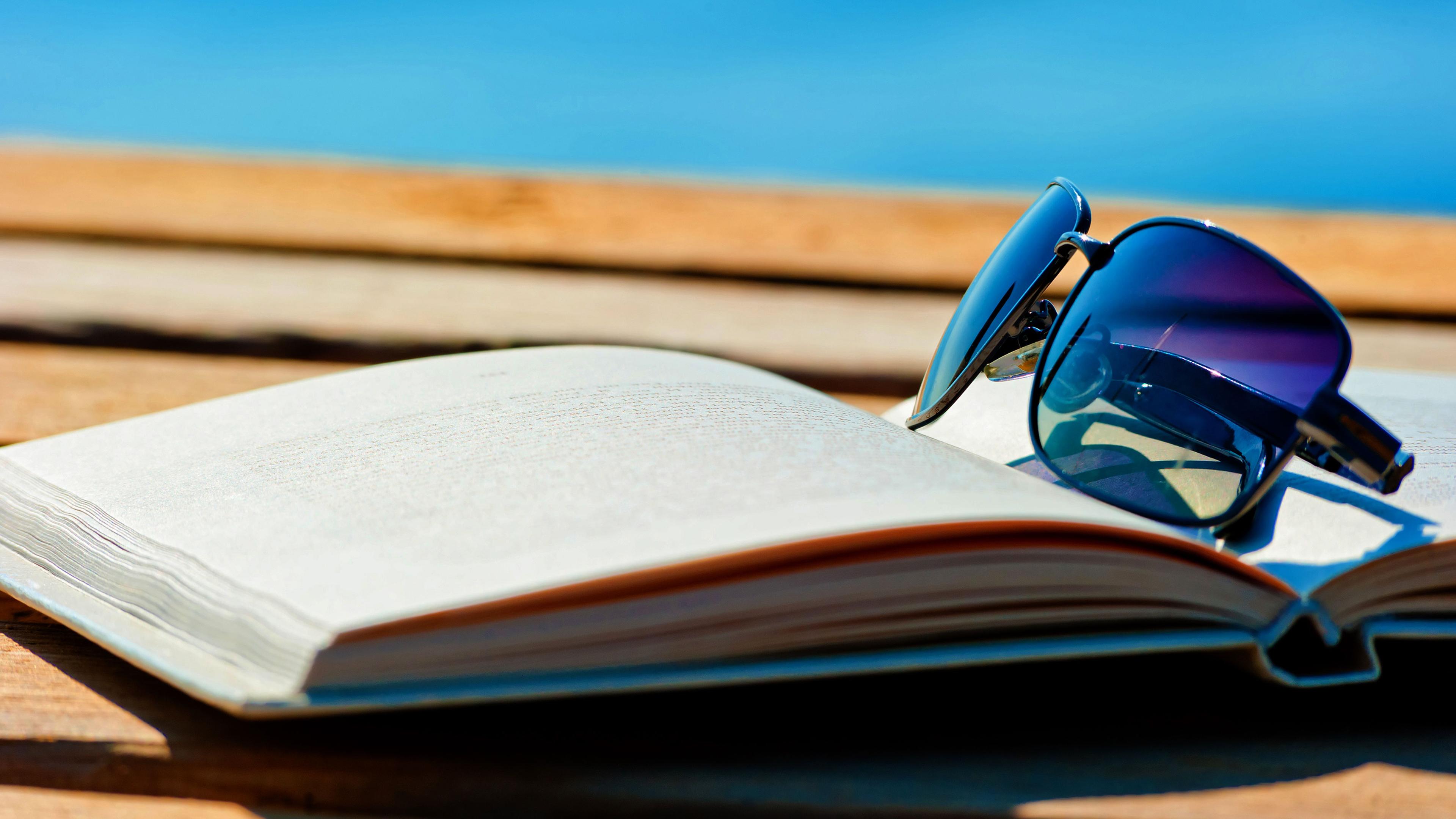 Ein aufgeschlagenes Buch mit einer daraufliegenden Sonnenbrille liegt auf einer Holzterrasse. Im Hintergrund ist das blaue Meer zu sehen.