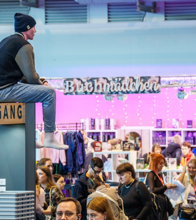 Ein Mann sitzt oben auf einer Stellwand zwischen Buchmessen-Gängen und Ständen über dem Menschengewusel.