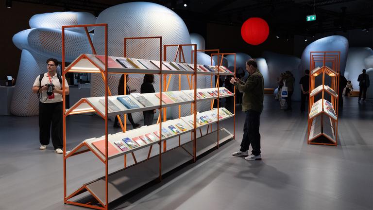 In einer abgedunkelten Halle steht ein filigranes Bücherregal aus Holz, vor dem ein Mann ein Buch liest. Im Hintergrund sind weitere Besucher zu sehen und runde futuristisch anmutende Objekte sowie eine rot leuchtende Kugel.
