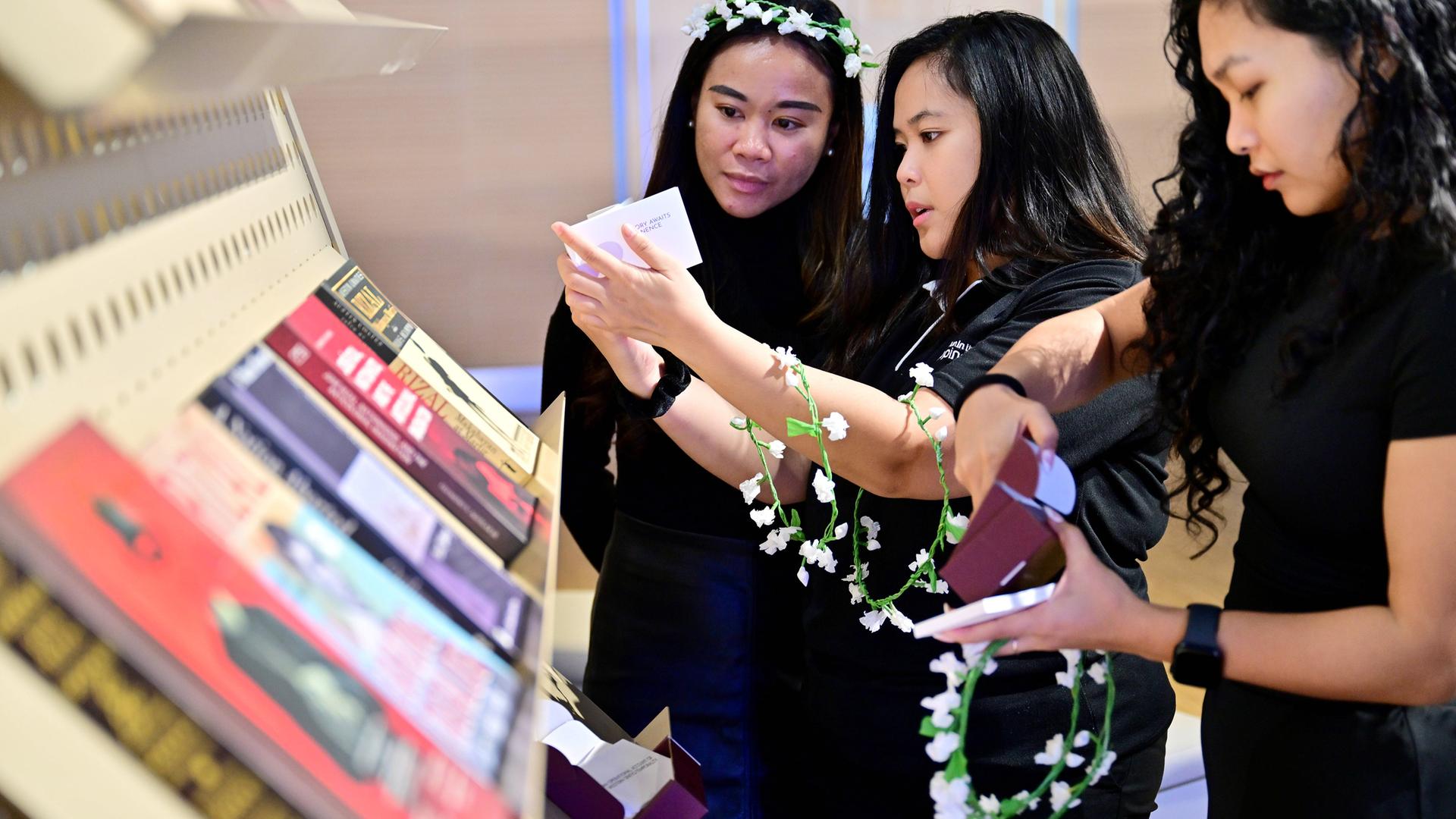 Drei junge asiatisch aussehnde Frauen halten schmale Blumenkränze in den Händen und betrachten ausgelegte Bücher an einem Stand.