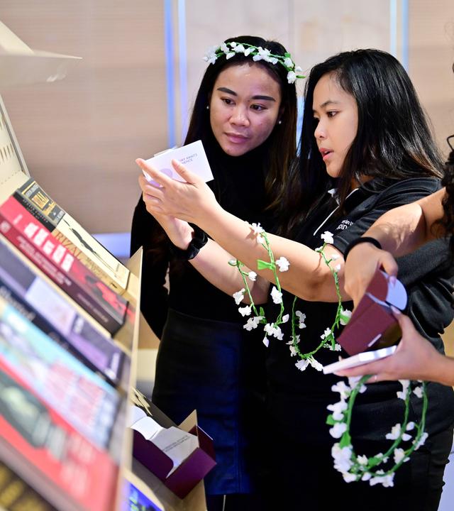 Drei junge asiatisch aussehnde Frauen halten schmale Blumenkränze in den Händen und betrachten ausgelegte Bücher an einem Stand.