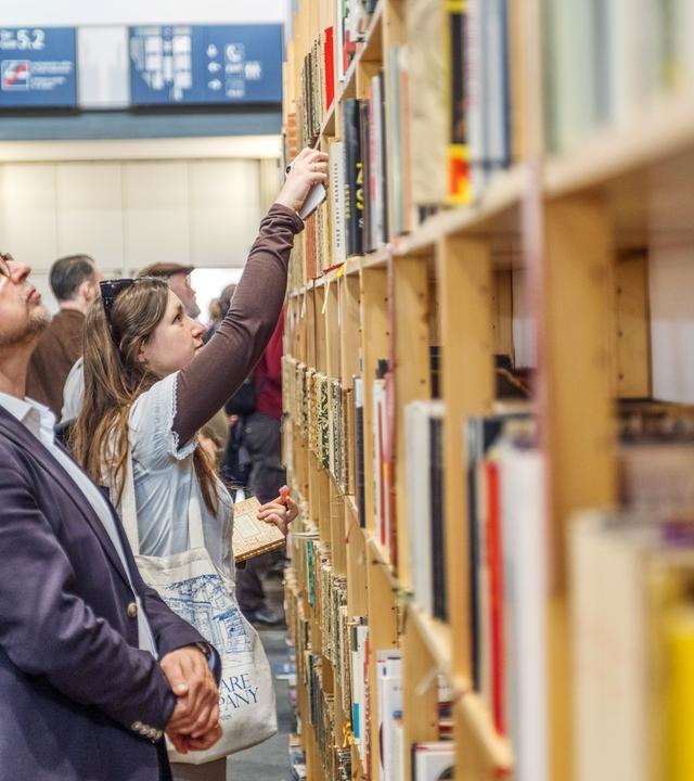 Vor einem großen Bücherregal stehen Menschen und betrachten die Buchrücken, oder ziehen Bücher heraus.
