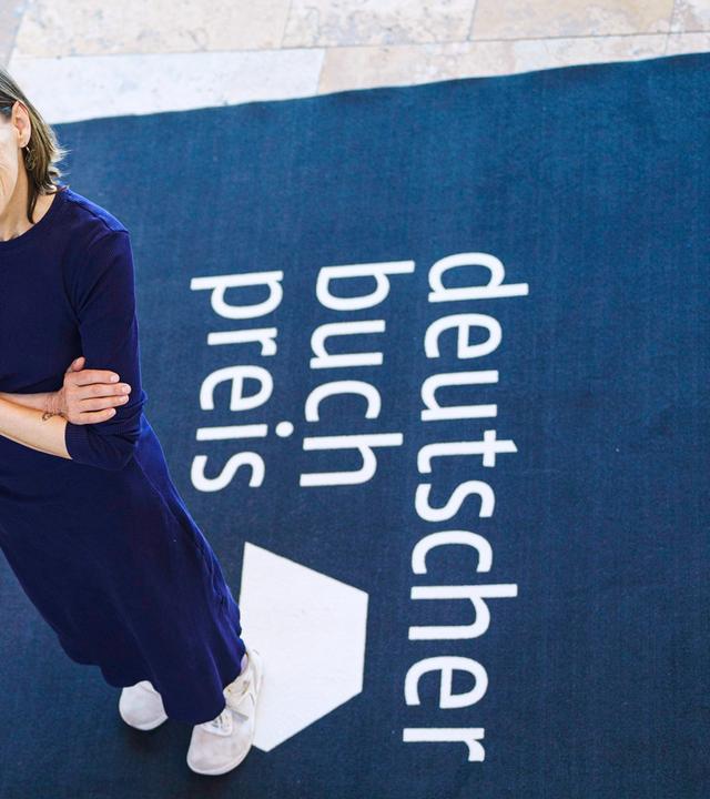 Eine Frau steht auf einem Teppich, auf dem deutscher buch preis steht und schaut nach oben in die Kamera.