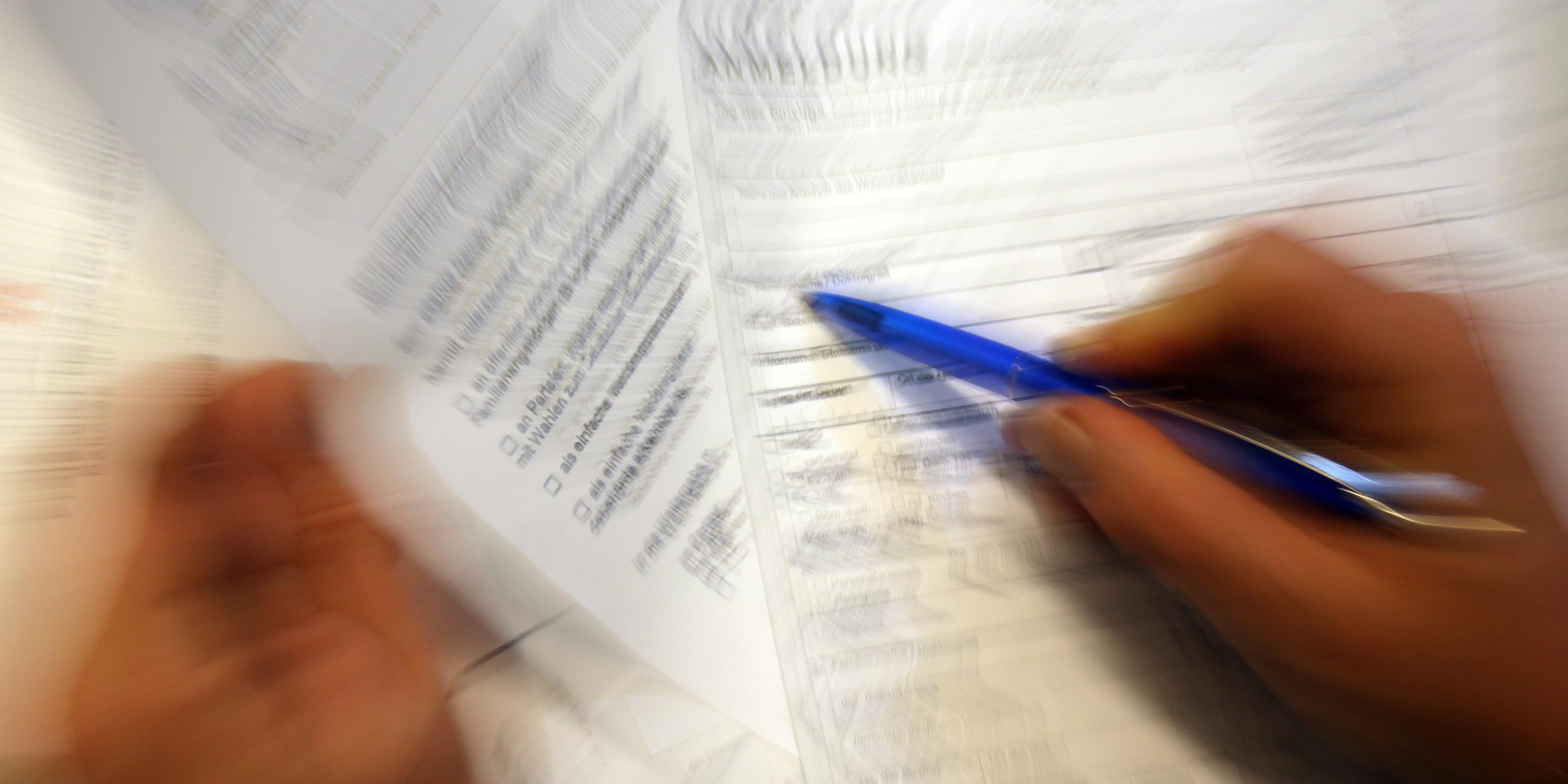Auf dem Bild sieht man ein verschwommenes Antragsformular. Eine Person setzt den Stift an, um das Blatt auszufüllen.