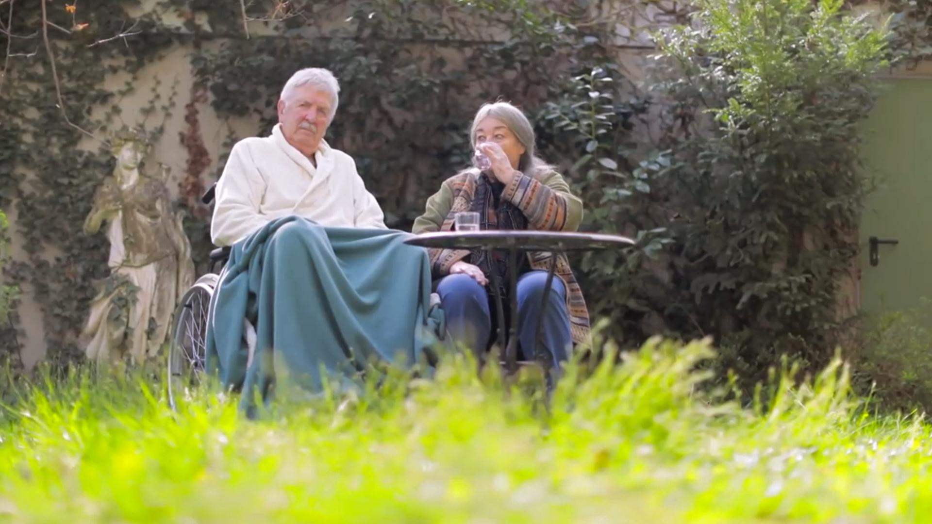 Szene aus "Der heutige Tag. Ein Stundenbuch der Liebe" von Helga Schubert: Ein altes Ehepaar sitzt im Garten. Der Mann sitzt in einem Rollstuhl. Er trägt einen weißen Bademantel und seine Knie sind mit einer türkisen Decke bedeckt. Neben ihm sitzt eine Frau, die aus einem Glas Wasser trinkt. Im Vordergrund steht ein brauner Gartentisch.