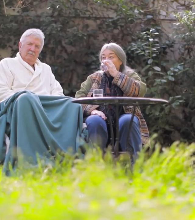 Szene aus "Der heutige Tag. Ein Stundenbuch der Liebe" von Helga Schubert: Ein altes Ehepaar sitzt im Garten. Der Mann sitzt in einem Rollstuhl. Er trägt einen weißen Bademantel und seine Knie sind mit einer türkisen Decke bedeckt. Neben ihm sitzt eine Frau, die aus einem Glas Wasser trinkt. Im Vordergrund steht ein brauner Gartentisch.