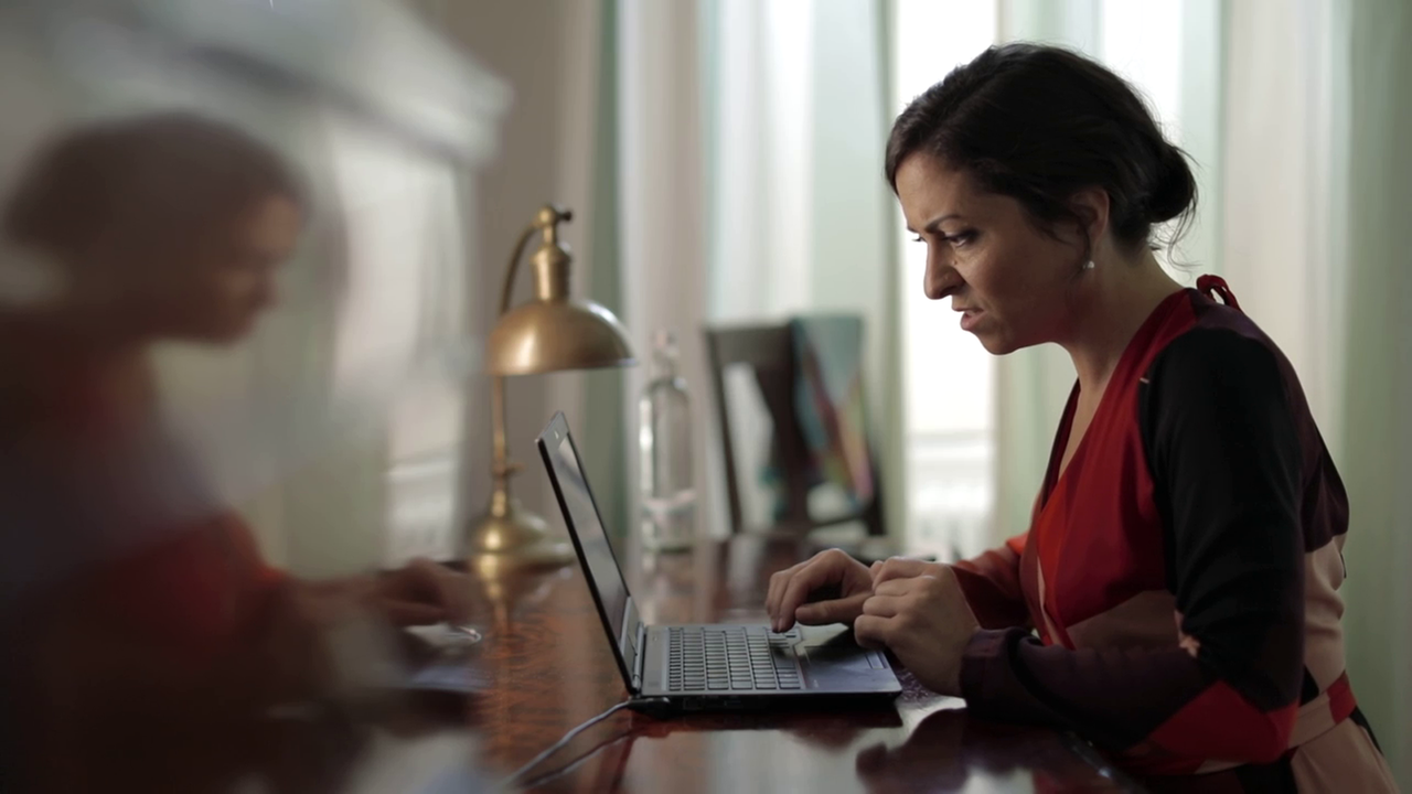 Szene aus "Der Pole" von John Maxwell Coetzee. Eine Frau sitzt an einem Tisch vor einem Laptop und liest. Ihr Gesichtsausdruck ist irritiert.
