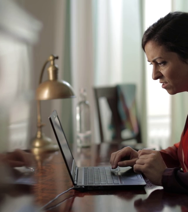 Szene aus "Der Pole" von John Maxwell Coetzee. Eine Frau sitzt an einem Tisch vor einem Laptop und liest. Ihr Gesichtsausdruck ist irritiert.