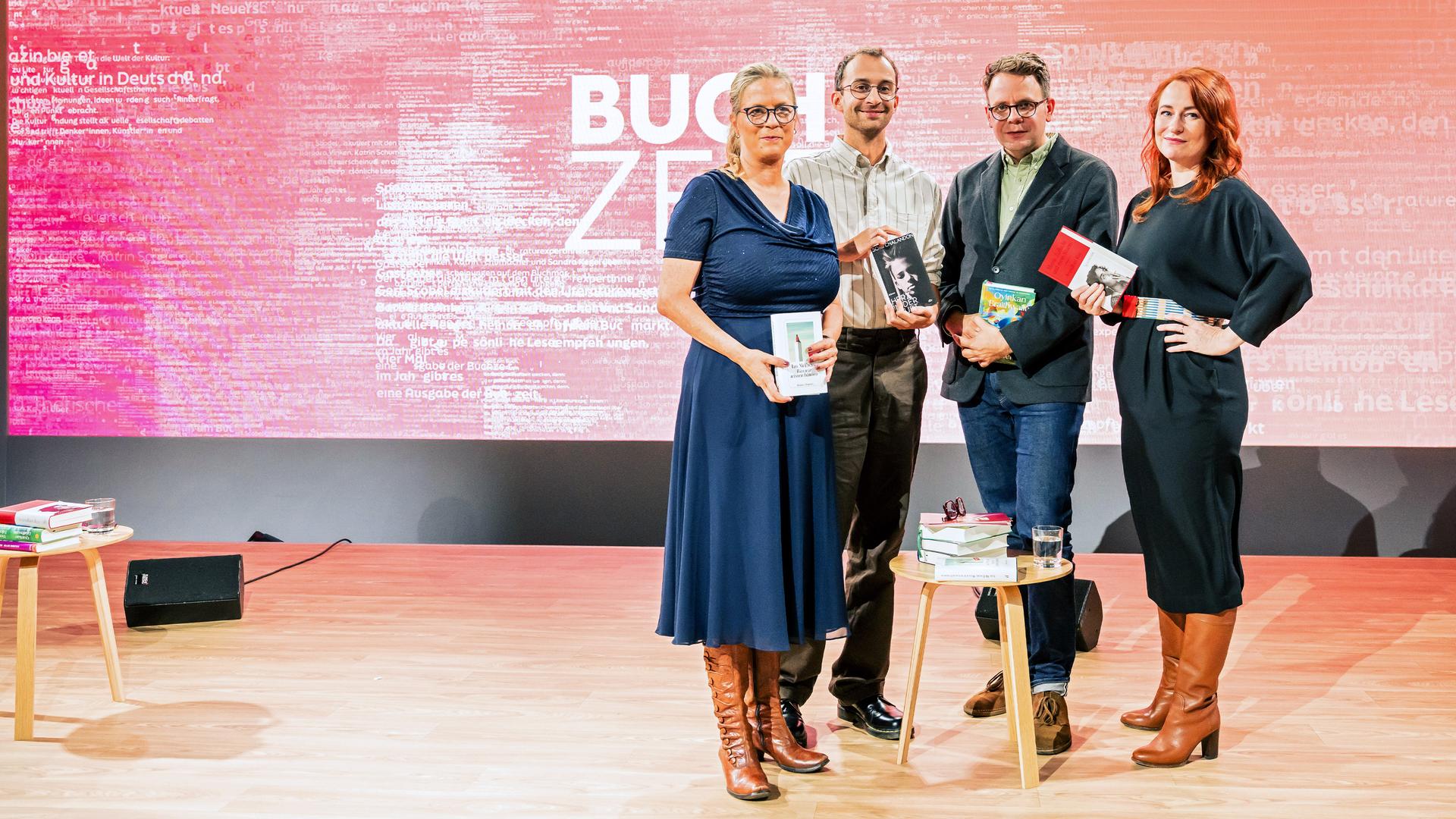 Vier Personen stehen auf einer Bühne mit pink-rotem Hintergrund, auf dem groß „BUCHZEIT“ zu lesen ist. Jede Person hält ein Buch in der Hand. Vor ihnen steh ein kleiner Tische mit Bücherstapeln. Auf der linken Seite steht ein zweiter kleiner Tisch mit einem Bücherstapel und einem Glas Wasser. Links neben einem Tisch steht ein Lautsprecher und der Bühnenboden ist aus Holz.  