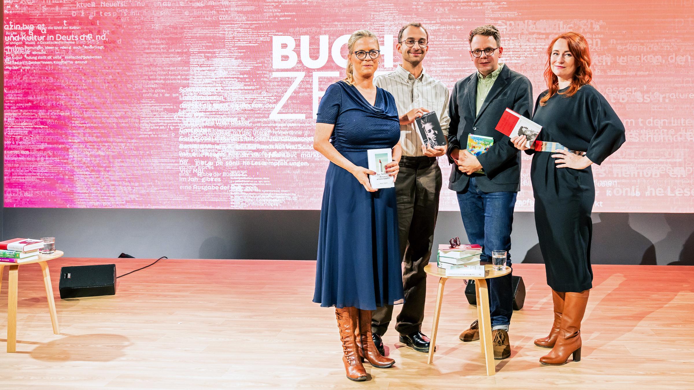 Vier Personen stehen auf einer Bühne mit pink-rotem Hintergrund, auf dem groß „BUCHZEIT“ zu lesen ist. Jede Person hält ein Buch in der Hand. Vor ihnen steh ein kleiner Tische mit Bücherstapeln. Auf der linken Seite steht ein zweiter kleiner Tisch mit einem Bücherstapel und einem Glas Wasser. Links neben einem Tisch steht ein Lautsprecher und der Bühnenboden ist aus Holz.  