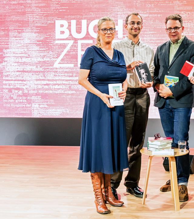 Vier Personen stehen auf einer Bühne mit pink-rotem Hintergrund, auf dem groß „BUCHZEIT“ zu lesen ist. Jede Person hält ein Buch in der Hand. Vor ihnen steh ein kleiner Tische mit Bücherstapeln. Auf der linken Seite steht ein zweiter kleiner Tisch mit einem Bücherstapel und einem Glas Wasser. Links neben einem Tisch steht ein Lautsprecher und der Bühnenboden ist aus Holz.  