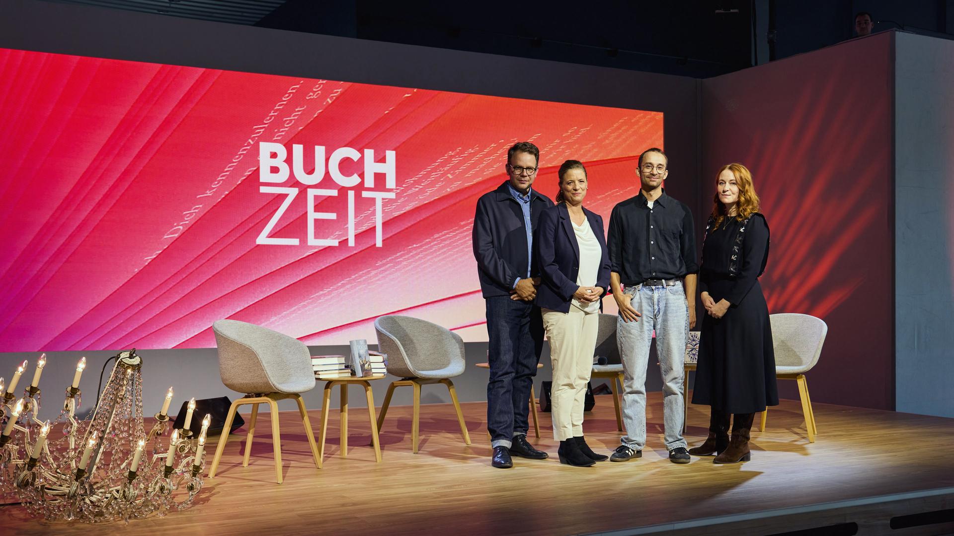 Gruppenfoto auf der Bühne der Literatursendung „Buchzeit“: Vier Personen posieren vor einer rot gestalteten Hintergrundwand mit Sendungslogo. Im Vordergrund stehen Stühle und Bücher.