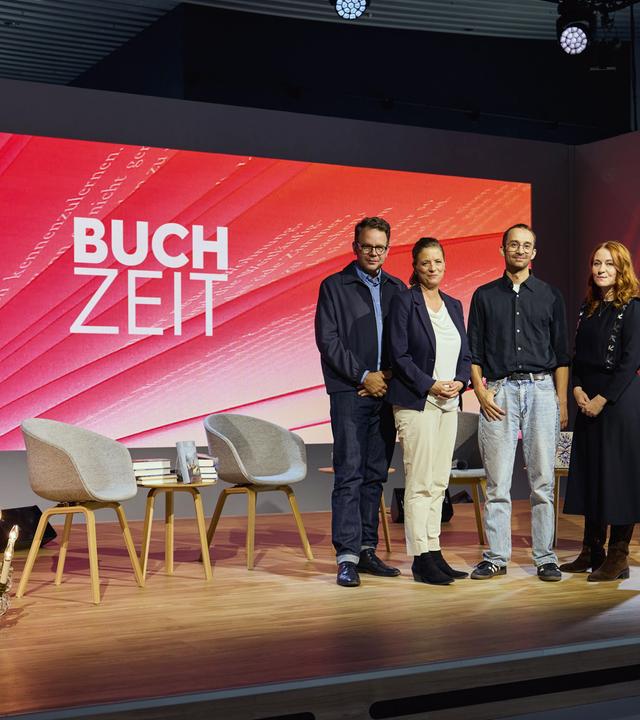 Gruppenfoto auf der Bühne der Literatursendung „Buchzeit“: Vier Personen posieren vor einer rot gestalteten Hintergrundwand mit Sendungslogo. Im Vordergrund stehen Stühle und Bücher.