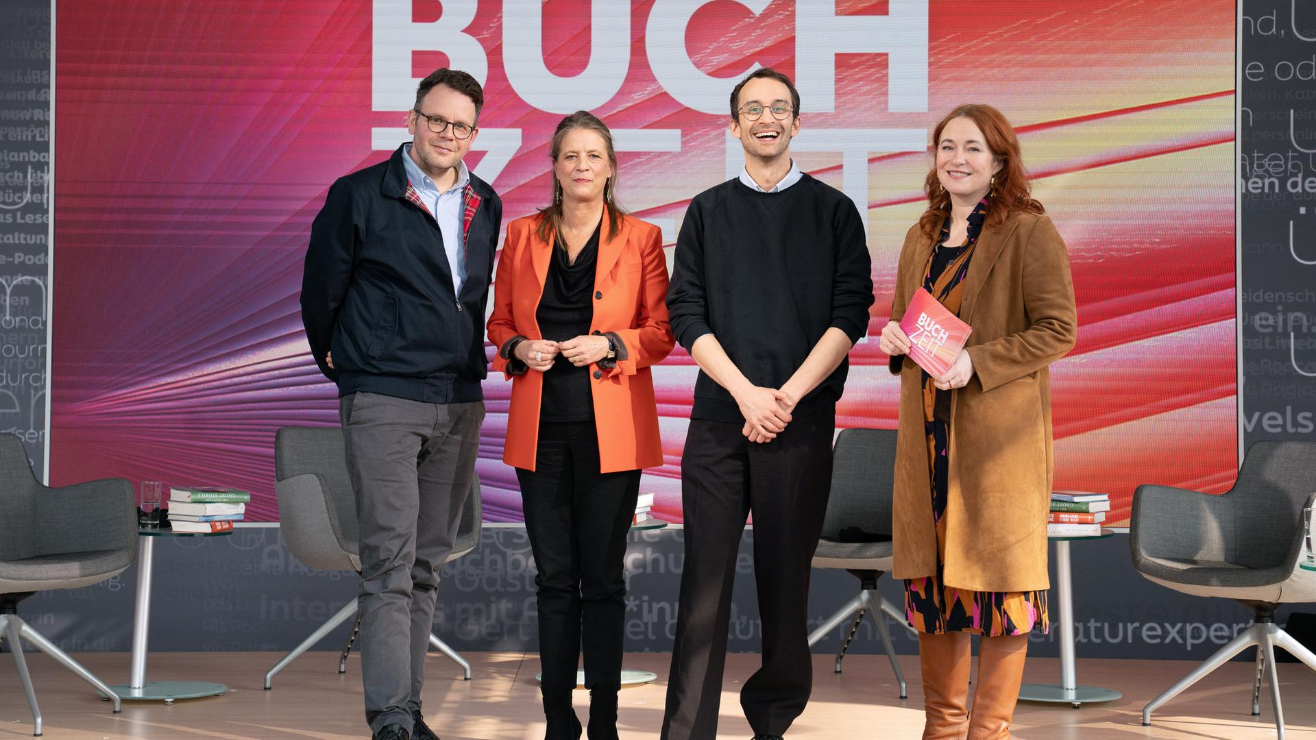 Gruppenfoto auf einer Studiobühne der Sendung „Buchzeit“. Vier Personen stehen auf einer beleuchteten Bühne mit dem Schriftzug „BUCHZEIT“ im Hintergrund; umgeben sind sie von mehreren Sesseln und Büchern als Teil des Bühnenbilds. 