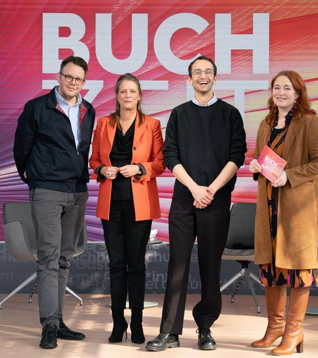 Gruppenfoto auf einer Studiobühne der Sendung „Buchzeit“. Vier Personen stehen auf einer beleuchteten Bühne mit dem Schriftzug „BUCHZEIT“ im Hintergrund; umgeben sind sie von mehreren Sesseln und Büchern als Teil des Bühnenbilds. 