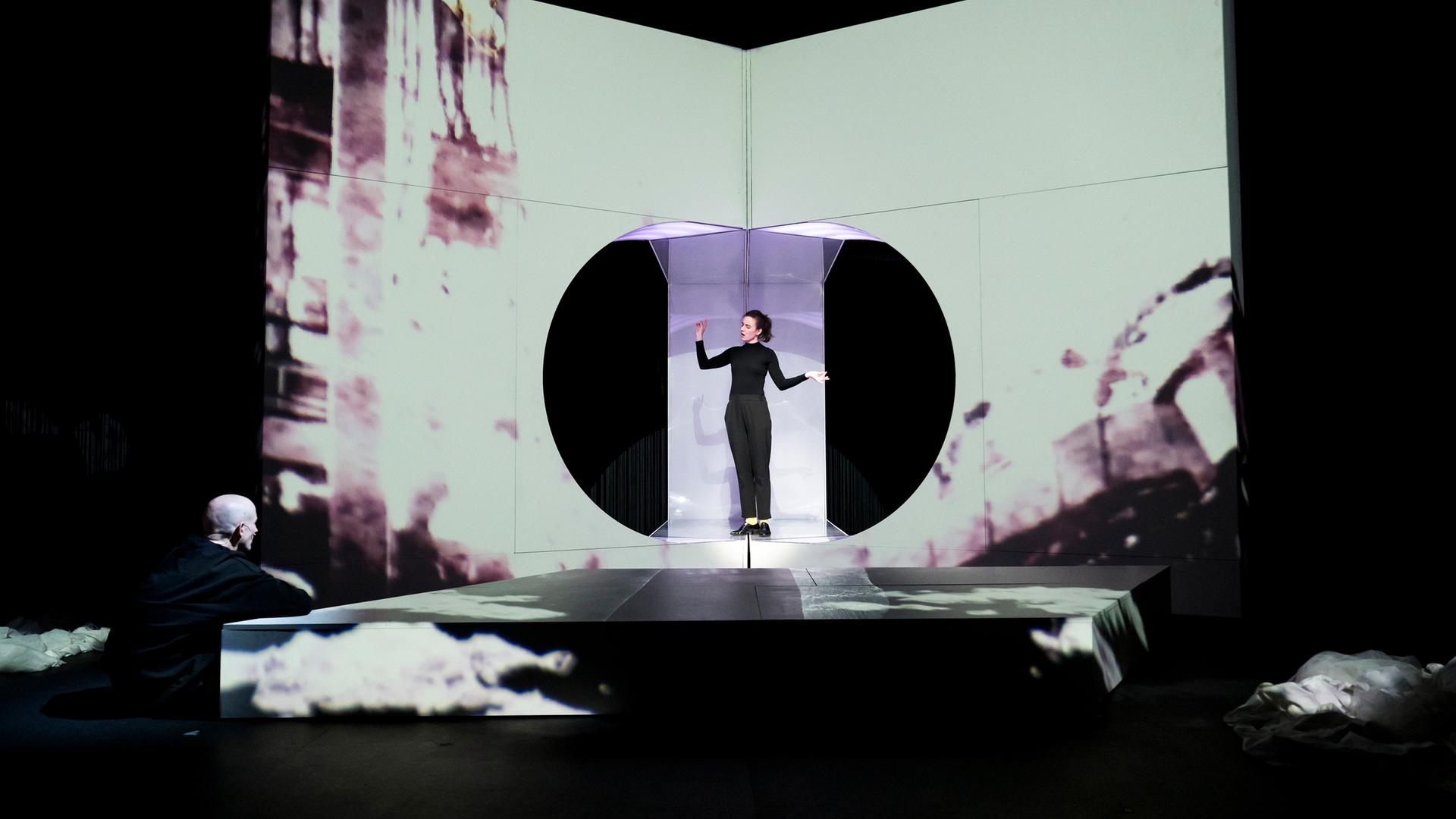 : Eine Wand mit einer sehr hellen Projektion, in der Mitte eine kreisrunde Öffnung, in der eine schwarzgekleidete, singende Frau steht. Vor ihr ein weißes, niedriges Podest. Links vorne sitzt ein glatzköpfiger Mann am Boden und sieht ihr zu, die Arme auf das Podest abgelegt.