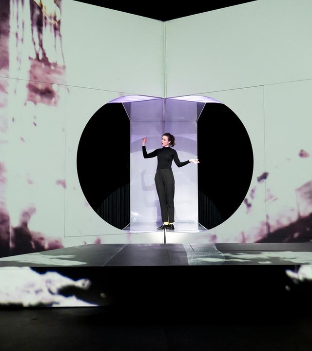 : Eine Wand mit einer sehr hellen Projektion, in der Mitte eine kreisrunde Öffnung, in der eine schwarzgekleidete, singende Frau steht. Vor ihr ein weißes, niedriges Podest. Links vorne sitzt ein glatzköpfiger Mann am Boden und sieht ihr zu, die Arme auf das Podest abgelegt.