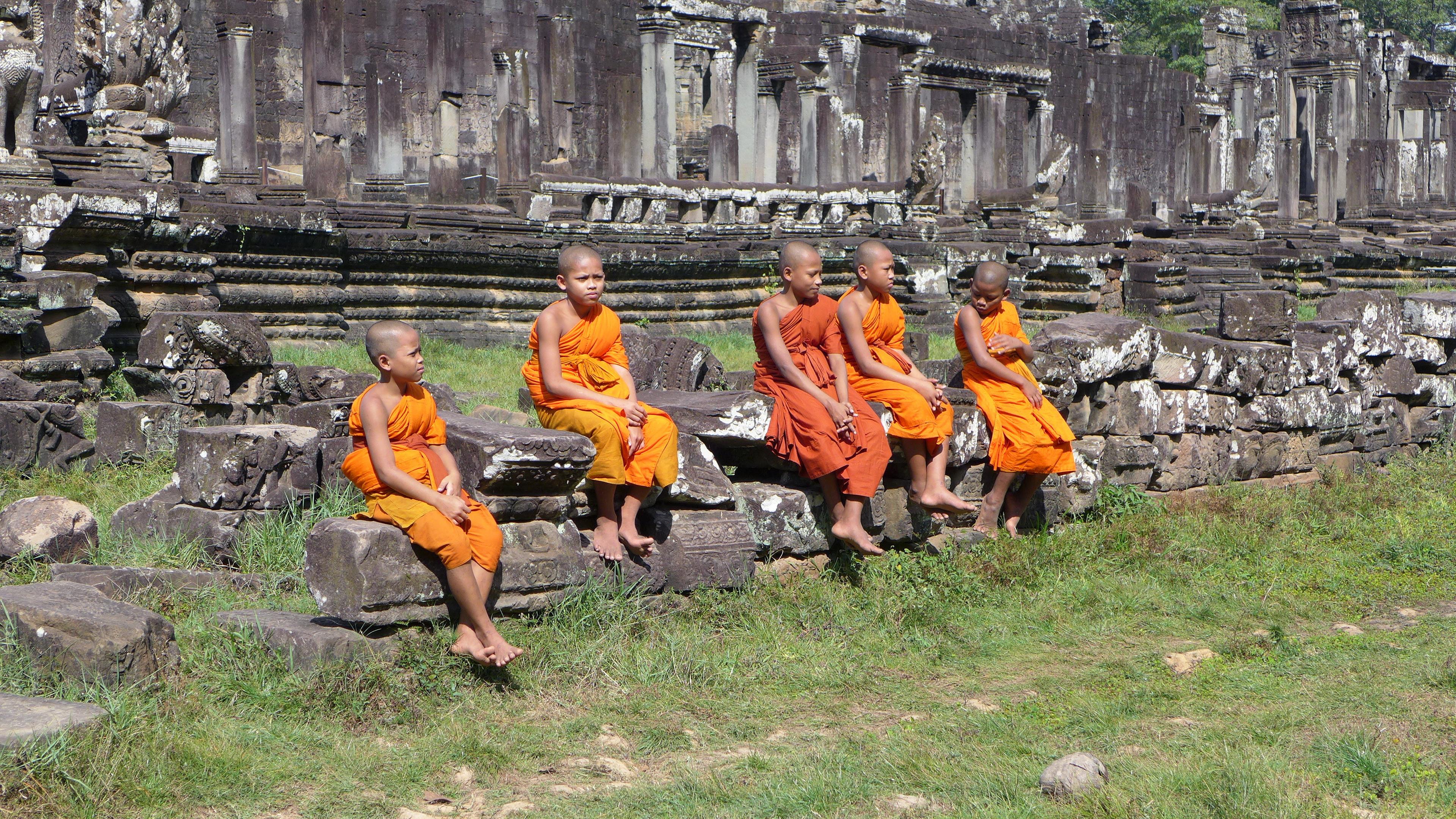 Eine Gruppe junger buddhistischer Mönche in traditionellen, orangefarbenen Gewändern, sitzt auf Steinen vor Angkor Wat.