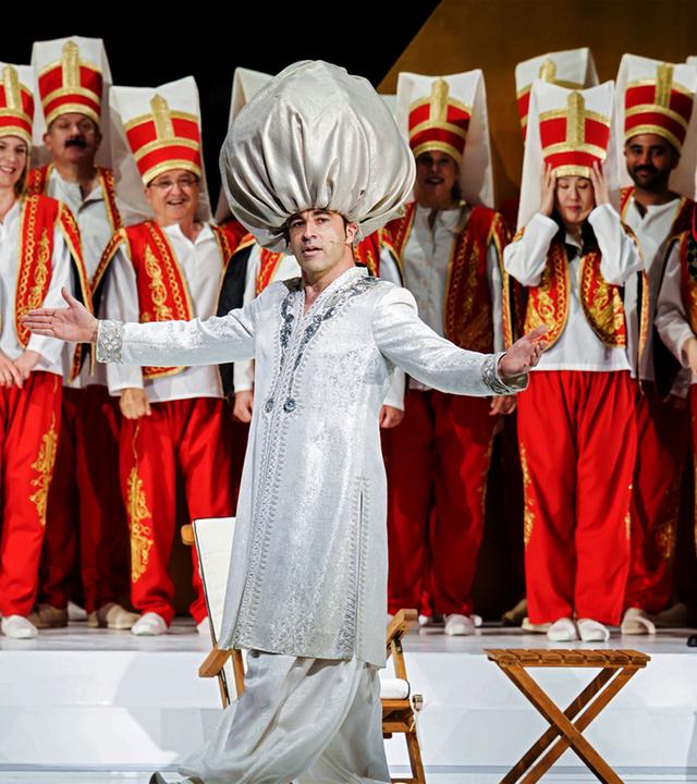 Eine Person in einem silberfarbenen Gewand mit großer Kopfbedeckung steht auf einer Theaterbühne vor einer Gruppe von Darstellerinnen und Darstellern in rot-weißen Kostümen. Im Vordergrund sitzt eine weitere Person neben einem kleinen Tisch.