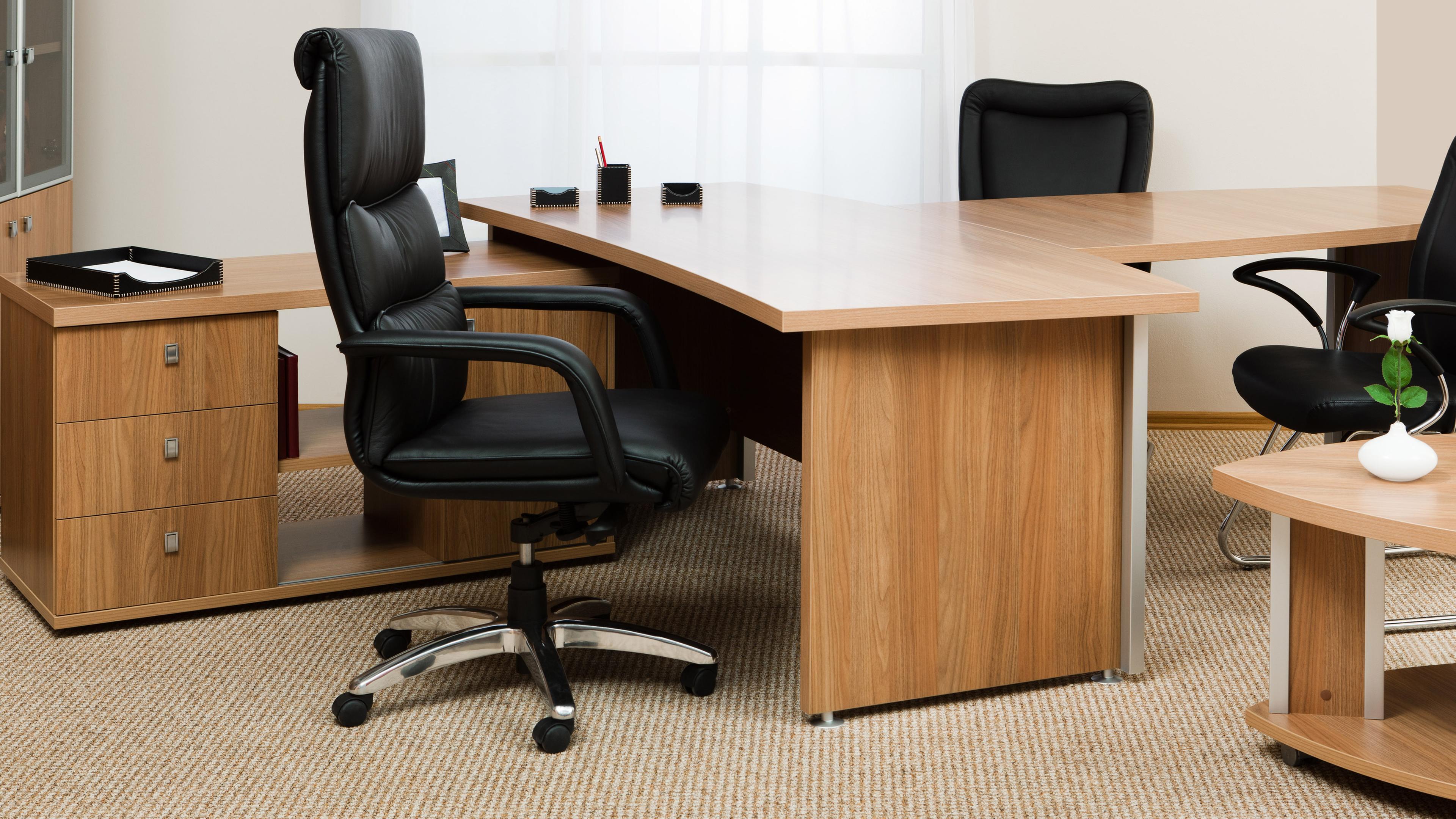 In einem hellen Büro befinden sich drei schwarze Bürostühle an einem Holzschreibtisch.