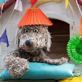 Hofhund Buffo liegt in seiner Hütte. Er trägt einen Lampenschirm auf dem Kopf und schaut nachdenklich.