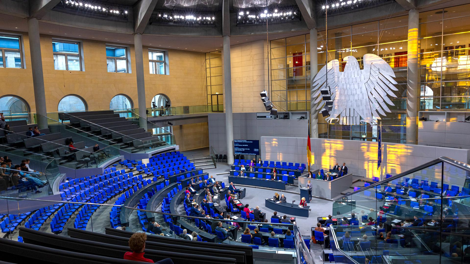 Ein großer hoher runder Saal, in dem ein riesiger Bundesadler an der Stirnseite hängt und Dutzende Reihen mit blauen Klappsühlen um ein Rednerpodest in der Mitte angeordnet sind.