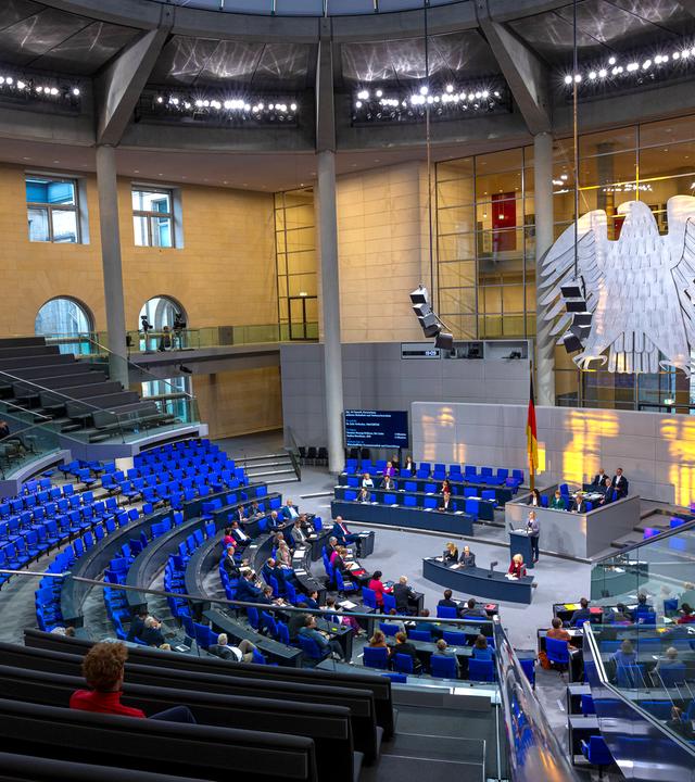 Ein großer hoher runder Saal, in dem ein riesiger Bundesadler an der Stirnseite hängt und Dutzende Reihen mit blauen Klappsühlen um ein Rednerpodest in der Mitte angeordnet sind.