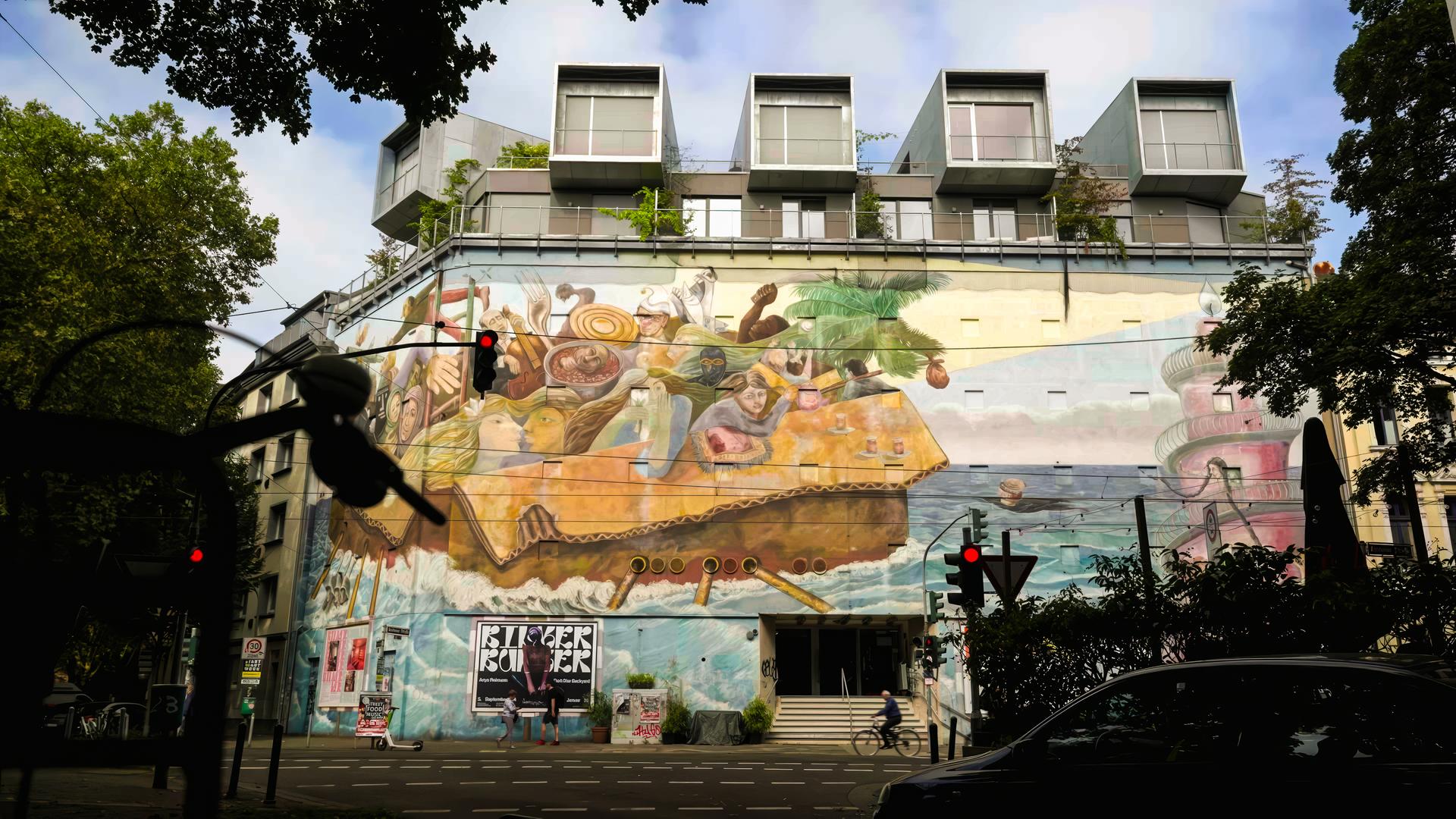 Eine hohe Gebäudefassade ist mit einem farbenfrohen Wandgemälde verziert. Das Mural zeigt Menschen und surreale Figuren in einem riesigen Pappkarton auf hoher See. 