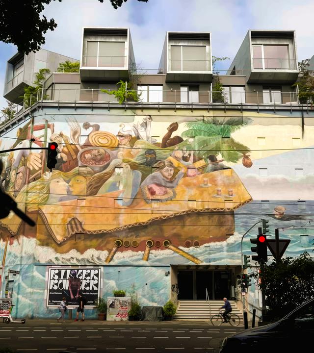 Eine hohe Gebäudefassade ist mit einem farbenfrohen Wandgemälde verziert. Das Mural zeigt Menschen und surreale Figuren in einem riesigen Pappkarton auf hoher See. 