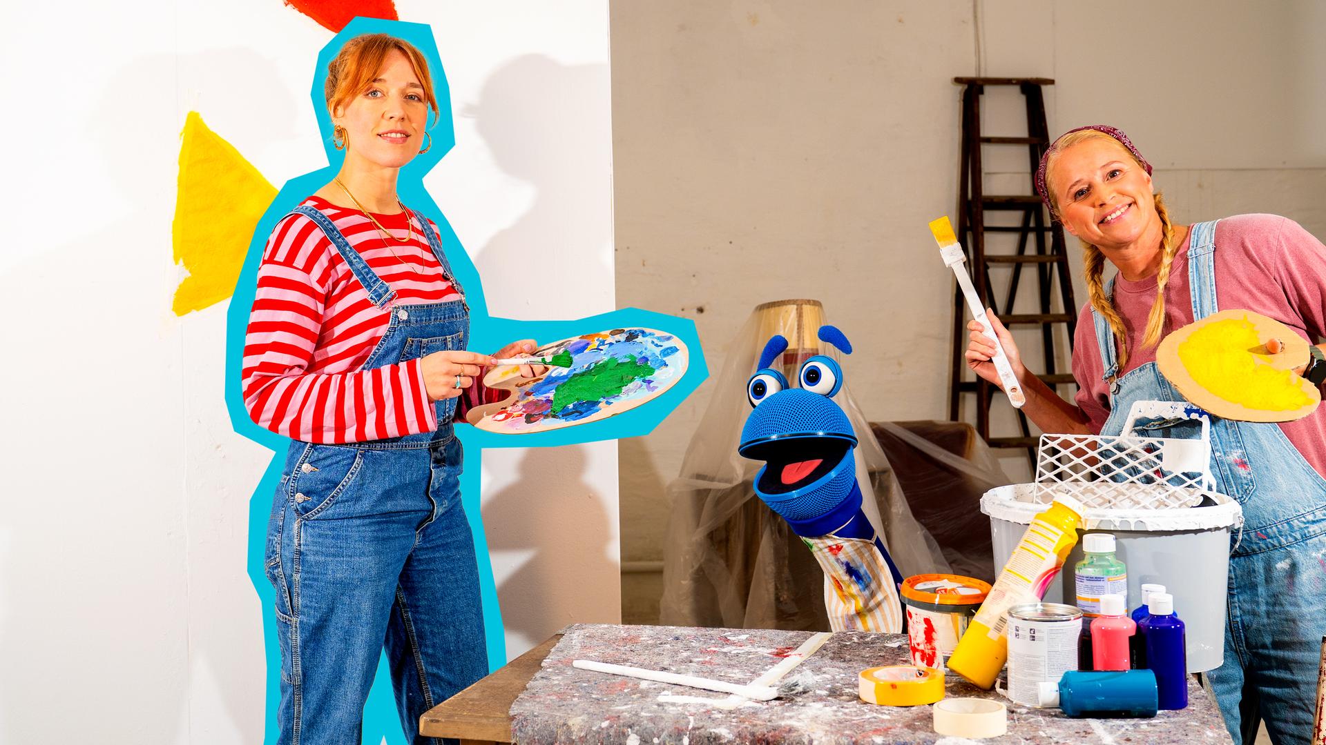 Auf dem Bild sind ANTE, SingDing und Singa zu sehen (v.l.n.r.). ANTE und Singa halten jeweils einen Pinsel und eine Farbpalette in der Hand - beide tragen außerdem eine Latzhose. Vor SingDing befindet sich ein Tisch auf dem Farbflaschen und ein Eimer steht. 