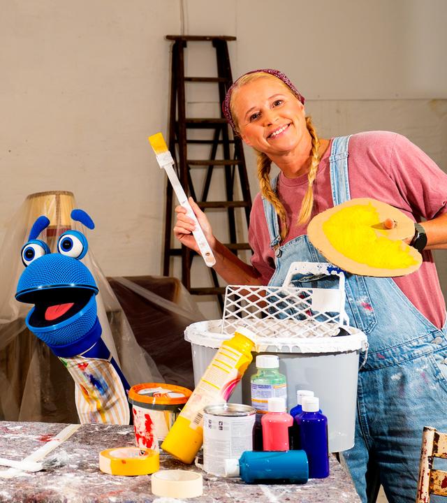 Auf dem Bild sind ANTE, SingDing und Singa zu sehen (v.l.n.r.). ANTE und Singa halten jeweils einen Pinsel und eine Farbpalette in der Hand - beide tragen außerdem eine Latzhose. Vor SingDing befindet sich ein Tisch auf dem Farbflaschen und ein Eimer steht. 