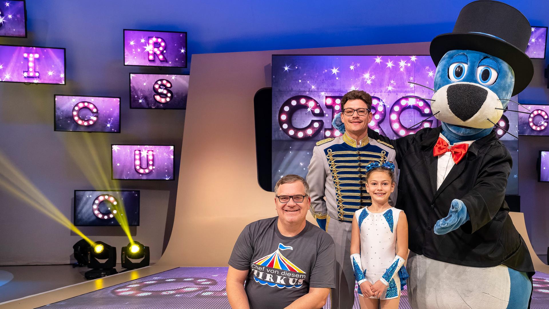 Simone Stey sitz im Spagat auf dem Studioboden. Zirkusdirektor Martin Stey steht dahinter. Eingerahmt werden sie von Elton und Piet auf der anderen Seite.