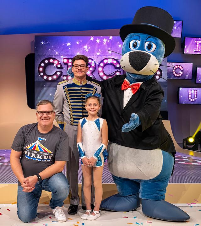 Simone Stey sitz im Spagat auf dem Studioboden. Zirkusdirektor Martin Stey steht dahinter. Eingerahmt werden sie von Elton und Piet auf der anderen Seite.