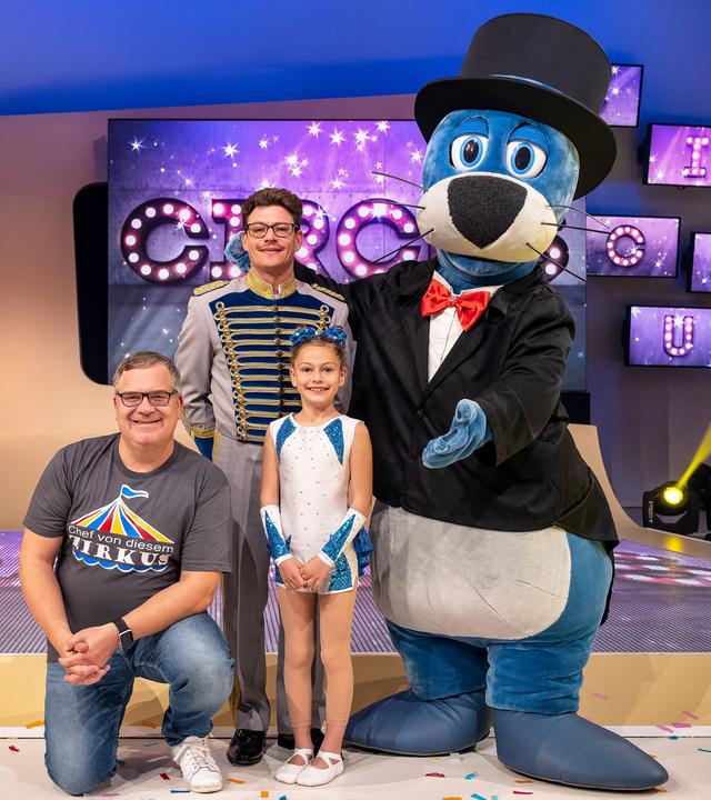 Simone Stey sitz im Spagat auf dem Studioboden. Zirkusdirektor Martin Stey steht dahinter. Eingerahmt werden sie von Elton und Piet auf der anderen Seite.