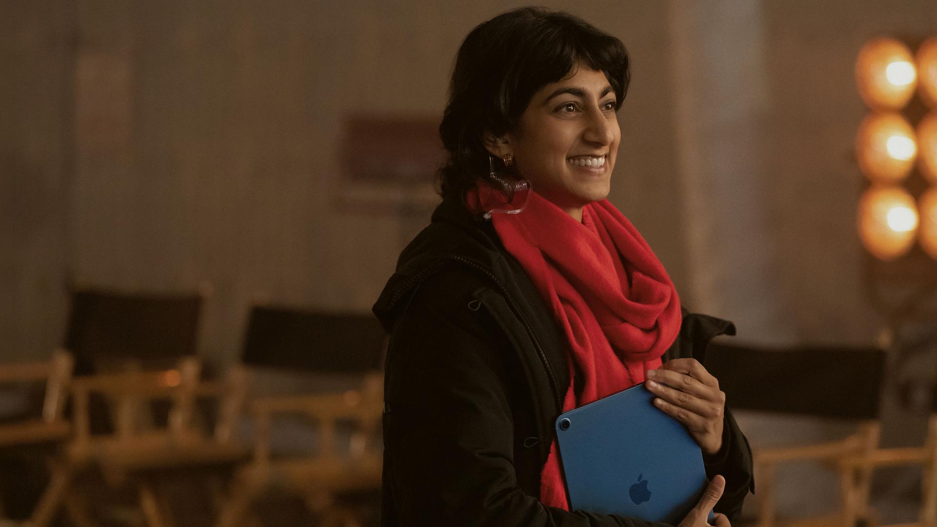 "Bupkis - Alles geht schief": Monica (Sunita Mani) steht am Filmset. Im Hintergrund stehen aufgereiht viele Stühle und die Scheinwerfer für den Dreh leuchten. Monika hält ein Notebook in der Hand, ihr roter Schal strahlt und sie schaut lächelnd zur Seite.