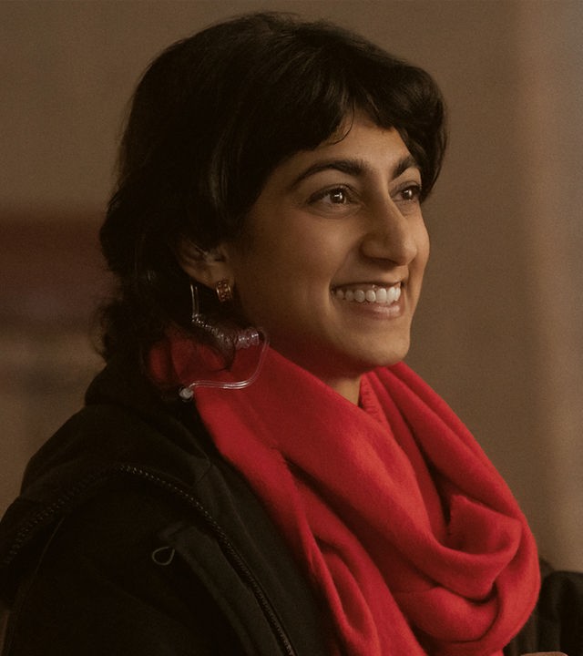 "Bupkis - Alles geht schief": Monica (Sunita Mani) steht am Filmset. Im Hintergrund stehen aufgereiht viele Stühle und die Scheinwerfer für den Dreh leuchten. Monika hält ein Notebook in der Hand, ihr roter Schal strahlt und sie schaut lächelnd zur Seite.