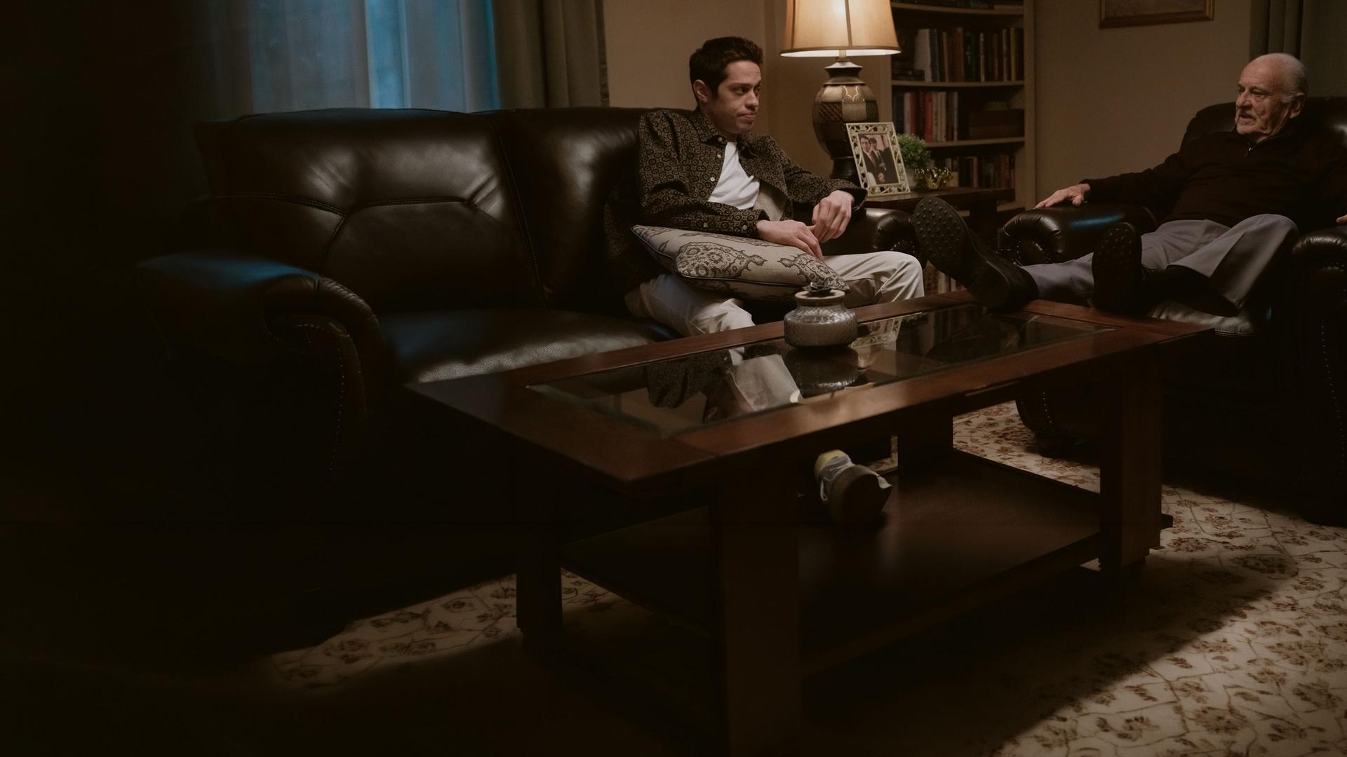 "Bupkis - Die Wandlung": Pete (Pete Davidson) sitzt auf einem Ledersofa. Auf seinem Schoß liegt ein Kissen. Er schaut Opa Joe (Joe Pesci) an, der im Ledersessel sitzt und mit Pete redet. Beide wirken sehr ernst. Der Raum ist dunkel, es leuchtet nur eine Tischlampe und die Stimmung wirkt bedrückend.