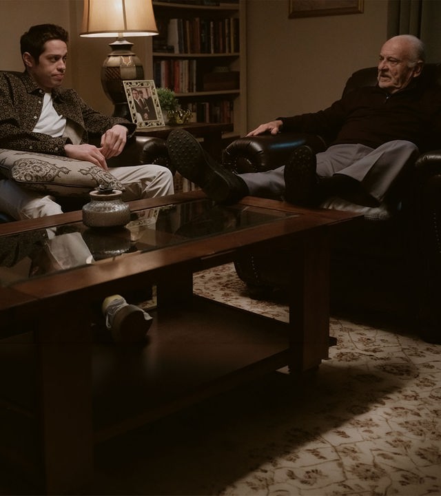 "Bupkis - Die Wandlung": Pete (Pete Davidson) sitzt auf einem Ledersofa. Auf seinem Schoß liegt ein Kissen. Er schaut Opa Joe (Joe Pesci) an, der im Ledersessel sitzt und mit Pete redet. Beide wirken sehr ernst. Der Raum ist dunkel, es leuchtet nur eine Tischlampe und die Stimmung wirkt bedrückend.